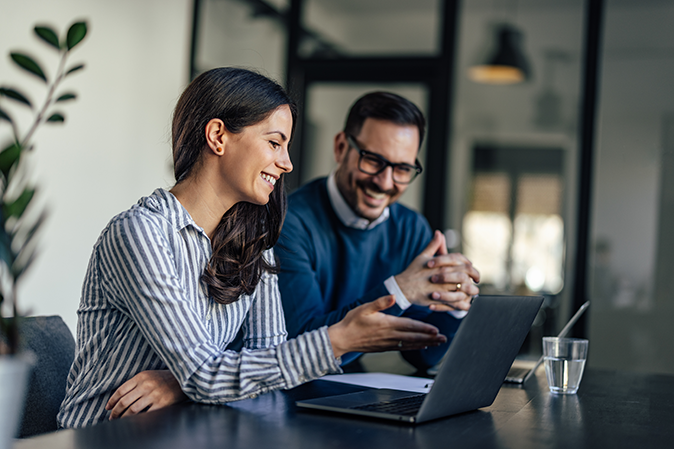 A young woman and a man smiling as they work together on a laptop in an office