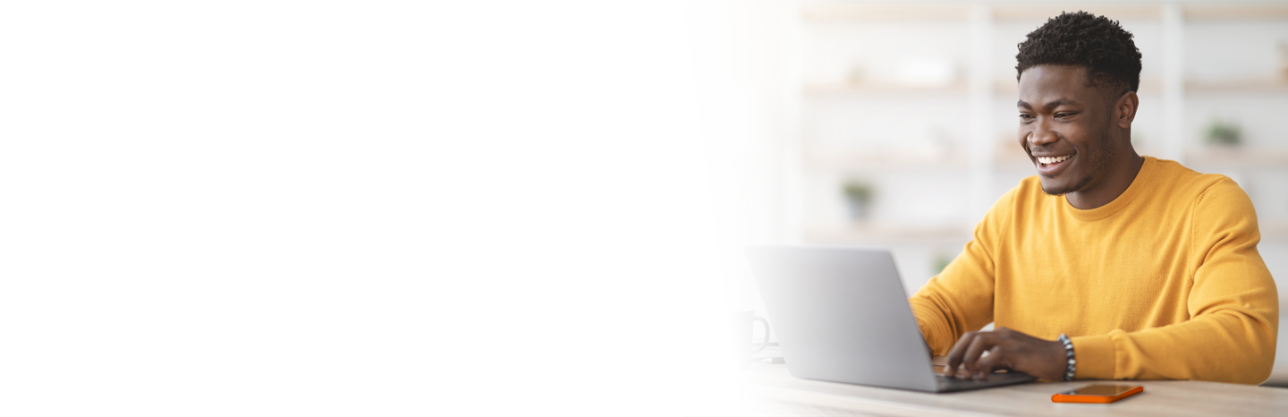 Young man smiling while typing on a laptop with his mobile device nearby