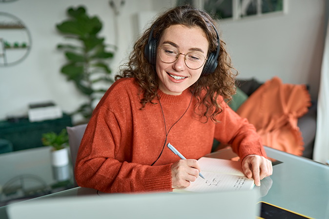 Young woman wearing headphones while taking notes watching a video on computer